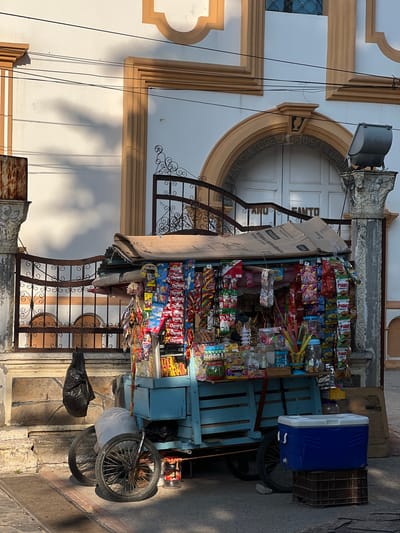 A candy cart is parked in front of a building in what is presumably La Esperanza, Honduras, evidenced by the text on the cart. The cart is laden with assorted candies and snacks, arranged in rows for display. A blue cooler sits atop the cart, beside a dark-colored crate. Black trash bags are visible beside the cart's wheels, which are on a paved surface. The building's architecture features a large arched window or door, ornate trim, and metal security bars. Electrical wires are strung across the facade. The scene is captured in daylight, with shadows indicating the direction of the sunlight.