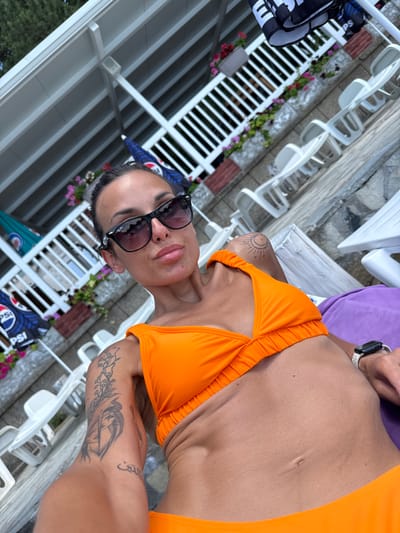 A woman is lying down on a lounge chair with a purple towel. She is wearing an orange bikini and sunglasses. Her hair is dark brown and pulled back. She has tattoos on her arm and leg. The background shows white lounge chairs, some umbrellas, and some plants. The scene appears to be outdoors, possibly at a resort or by a pool.