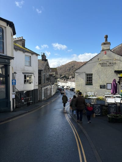 The image depicts a street scene in Ambleside, United Kingdom. The view is looking down a wet asphalt road with double yellow lines, sloping gently downhill.

On the immediate right, the prominent building is "THE PRIEST HOLE RESTAURANT," a light-colored, textured building with a pitched roof, a decorative chimney, and gold wall-mounted lights. Its facade features a large window with a planter box. An outdoor dining area is set up with multiple white tables and chairs, flanked by planter boxes and a folded purple umbrella. Several dark sandwich board signs display "EVERY TUESDAY FISH NIGHT" and "EVERY THURSDAY 5PM STEAK NIGHT."

On the left, a row of traditional stone and stucco buildings lines the street. The first building on the left has a dark lower facade and a white upper section with a large arched window. A blue one-way arrow sign is affixed to a pole on the sidewalk. Adjacent to it is a white stucco building with a bay window and a roof partially covered in moss. Further down the street, buildings constructed from dark stone and slate roofs are visible, some with signs, including "AMBLESIDE LAUNDRY."

Six individuals are walking away from the camera in the foreground and midground. Four people form a group on the right side of the road, consisting of two adults and two children. One adult, wearing a brown jacket, holds hands with a child in a red and blue jacket carrying a small paper bag. Another adult in a green jacket and a further adult in a brown jacket are also part of this group. Two additional individuals are visible further down the street on the left, also walking away.

The street continues into the distance, with several parked cars visible along both sides. In the background, rolling hills or fells, partially covered with brown and green vegetation, rise under a bright blue sky dotted with scattered white clouds.