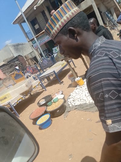 The scene depicts an outdoor commercial area in Omupo, Nigeria, under bright daylight. In the immediate foreground on the right, a dark-skinned male subject is visible in profile, facing left and slightly downwards. He wears a multi-striped, patterned cap and a dark shirt with horizontal stripes and geometric patterns. His shadow is cast on the reddish-brown dirt ground.
To the left and front of the main subject, several containers hold various food items for sale. These include multiple black plastic tubs and smaller colored bowls filled with grains, such as yellowish corn kernels and reddish-brown legumes (likely groundnuts/peanuts). Adjacent to these containers, a pile of white, irregularly shaped chunks (possibly dried cassava) rests on a dark ground cover, with a yellow bucket containing similar chunks beside it.
In the midground, several metal bed frames, some fitted with mattresses (one labeled "UNIFOAM"), are displayed. These frames feature varying designs and colors, including blue with ornate headboards. A wooden table structure is partially visible behind them.
The background features a two-story building with a light-colored facade and a corrugated metal roof. The building has a balcony with a black railing on the second floor. Signs, including a green banner, are visible on the ground floor, indicating commercial activity. To the left of this building, another structure with stacked materials is present. A tall, slender pole extends vertically into the clear blue sky from the left side of the frame. Another male subject wearing eyeglasses and a dark garment is partially visible behind the foreground individual, towards the right side of the frame. Faint outlines of bicycles or motorcycles are visible further in the background.