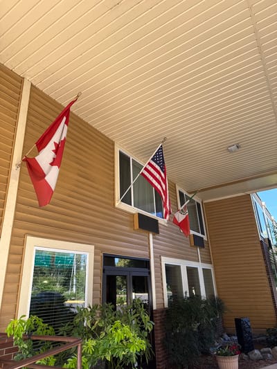 The image shows the exterior of a building with a tan, ribbed siding. Flags are displayed on the building. A Canadian flag is mounted on a pole near the bottom of the image, with the red maple leaf clearly visible. Above it, an American flag is also mounted on a pole. Further up, a Mexican flag is visible, hanging at an angle from a pole.
The building has several windows. Some windows have dark shutters. There is also greenery and plants around the building, with a potted plant containing red flowers visible on the right side. The sky is visible at the top, suggesting it is daytime. The scene appears to be a residential or commercial building, possibly a home or a business. The flags suggest a connection to Canada, the United States, and Mexico, indicating international relations or diverse occupancy. There is no visible text in the image.