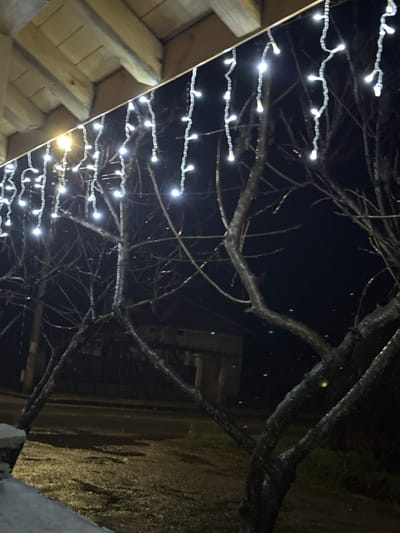 The image depicts a nighttime outdoor scene in Касапите, Bulgaria. Overhead, the underside of a wooden architectural structure, featuring light-toned exposed wooden beams and planks, is visible. Hanging from its edge are multiple strands of white LED icicle-style string lights, illuminating the immediate foreground. A bright yellow-orange light source, likely a porch light or streetlamp, is present on the far left, casting a localized glow. In the mid-ground, several bare-branched deciduous trees stand, their dark branches partially reflecting the light. The ground in the lower frame appears wet or damp, showing reflections from the lights, and consists of a mix of soil, sparse vegetation, and a darker, possibly paved, surface further back. A light-colored concrete or stone low wall or curb is visible in the bottom-left corner. The background is dark, with faint, indistinguishable outlines of distant structures or natural elements. No people are visible.