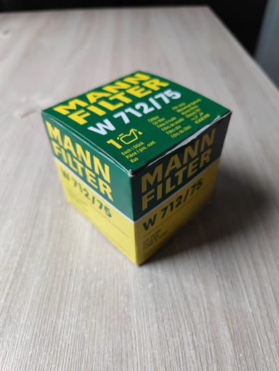 A MANN FILTER W 712/75 oil filter box is displayed on a light-colored wooden surface. The box is predominantly green and yellow, with "MANN FILTER" and "W 712/75" prominently printed in yellow and white respectively. The box also features various translations of "oil filter" in different languages and a small oil can graphic. The image is taken from a slightly elevated angle, showing the top and side of the box. There is no discernible connection to Nefteyugansk, Russia, or any people, actions, or interactions.