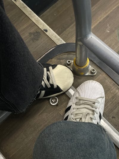 The image shows a close-up, overhead view of two pairs of feet, wearing Adidas Superstar sneakers, on a wooden floor. One person is wearing white Adidas Superstars with black stripes, and dark blue denim jeans. The other person is wearing black and white Adidas Superstars with white laces, and dark grey denim jeans. Both individuals appear to be seated or resting, as their feet are positioned on the floor, with one leg crossing over the other. A metal railing, possibly from a bus or train, runs diagonally across the top left of the frame. The floor is made of wooden planks, and there are several circular metal fixtures visible on the floor, likely part of the seating or structure of the vehicle. The overall lighting suggests an indoor setting, possibly during the day. There is no visible text in the image. The context suggests this scene is taking place in Yakovlevskoye, Russia.