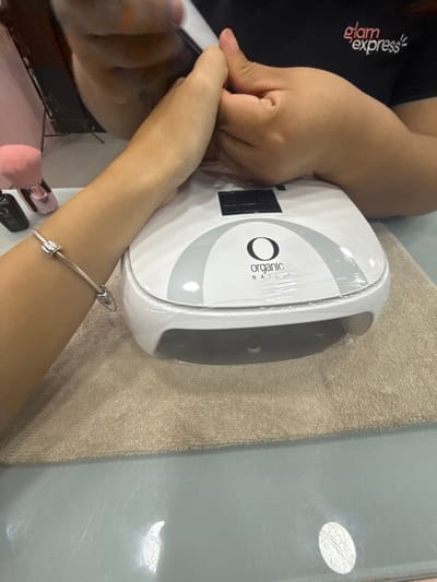 The image depicts a close-up view of a nail care service in progress at a "Glam Express" salon. A technician, wearing a black uniform shirt bearing the "glam express" logo, is actively filing the nails of a client's left hand. The client's left forearm, adorned with a silver bracelet featuring a square charm, is positioned on a beige terrycloth towel. Adjacent to the client's hand is a white "ORGANIC NAILS" branded UV/LED nail lamp, encased in protective clear plastic film. The service is occurring on a light blue-grey tabletop surface. Additional salon items, including a pink brush and a black nail polish bottle, are partially visible in the background. The specific address within Santa Catarina, Mexico, is not discernible from the image.
