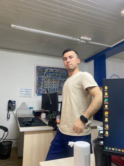 A man is standing in an office setting. He is wearing a light-colored t-shirt and dark blue shorts. On his left wrist, he has a black smartwatch. The man is standing next to a desk that has a computer monitor, keyboard, mouse, telephone, printer, and a roll of wet wipes. There is a large diagram or map on the wall behind the desk. The ceiling has fluorescent lighting fixtures. The man is looking towards the camera with a neutral expression. The overall impression is a typical office environment, likely in an industrial or administrative building. The specific location in the city of Yukhta, Russia, is not indicated by any visual cues within the image.