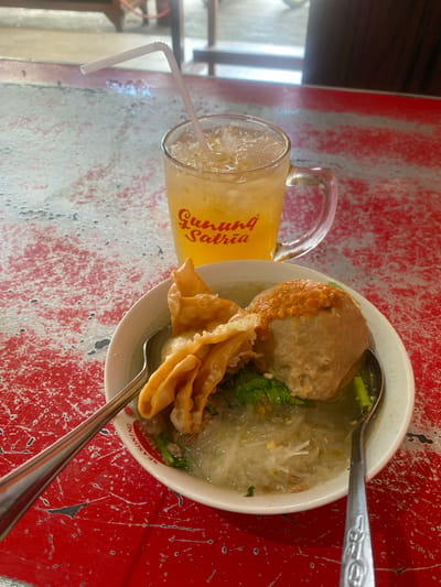 A close-up, eye-level shot shows a bowl of bakso (Indonesian meatball soup) and a glass of iced tea with a straw, placed on a textured red table. The bakso soup contains a large meatball topped with chili sauce, a few fried wontons, green vegetables, and thin rice noodles in a clear broth. The glass of iced tea is filled with ice cubes and has a bent white straw. The table has a distressed red painted surface with visible scratches and wear. In the background, blurry wooden furniture and a brightly lit outdoor area suggest a restaurant or eatery setting. The overall impression is of a casual meal enjoyed at an establishment, possibly in Sungai Sipai, Indonesia, given the "Gunung Satria" branding on the glass, which is a known business in the area.
