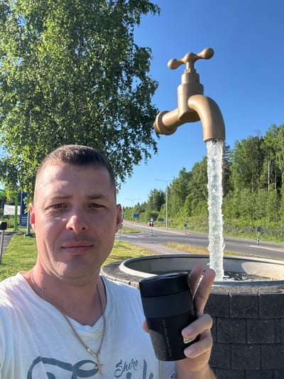 A man in a white t-shirt and a gold chain poses for a selfie in front of a large, golden faucet sculpture that is spraying water into a basin. The man is holding a black thermal cup and making a peace sign with his left hand. In the background, there is a road with some cars, trees, and a blue sky. This is a roadside attraction in Heinola, Finland, known as the "Tap Fountain" or "Vesihanasta" in Finnish. The sculpture is a popular spot for tourists to take pictures.