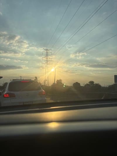 The image depicts a scene on a road in Tultitlán, Mexico, at sunset. The sun is partially obscured by the clouds, creating a halo effect. High-voltage power lines stretch across the sky. The back of a white Volkswagen car is visible, with its tail lights illuminated. The view is from inside another car. Trees line the side of the road. The sky is filled with clouds and the overall lighting indicates the end of the day.