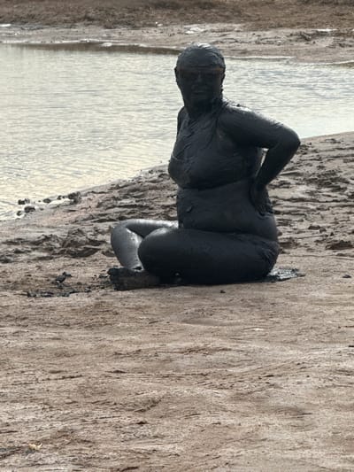 A person, covered head-to-toe in dark mud, sits on a muddy bank next to a body of water. The person is wearing sunglasses. The mud appears to be a thick, viscous substance. The surrounding environment is sandy and barren, with the water appearing murky. The scene is located in the Yamyyshevsk rural district of Kazakhstan.