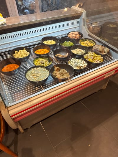 A display case holds a variety of prepared food items in black bowls. The offerings include various salads, vegetables, and saucy dishes, showcasing a range of colors and textures. Bowls are arranged in rows, with some containing creamy salads, while others feature bright orange or green components. The display case has a glass front and a metal grid inside.