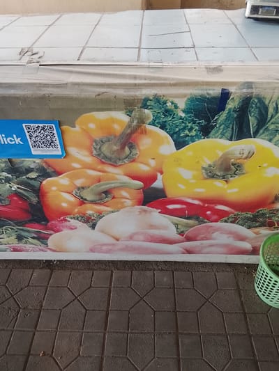 The image depicts a close-up, top-down view of a market stall in Sariosiyo, Uzbekistan. The foreground is dominated by patterned cobblestones, suggestive of an outdoor urban setting. Above the cobblestones, a large banner or tarp displays a vibrant image of various vegetables, primarily bell peppers in yellow and orange, along with red peppers and what appear to be radishes or onions. A blue sign with the word "lick" and a QR code is affixed to the banner on the left side. In the upper right corner, a small portion of a digital scale is visible, indicating a commercial transaction environment. A green plastic basket rests on the edge of the cobblestones on the right, partially obscuring the banner. The overall scene suggests a produce market or a food vendor's display.