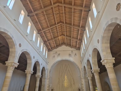 The image shows the interior of a church in Abu Gosh, Israel, featuring a high, wooden-beamed ceiling and stone arches supported by ornate columns. Sunlight streams in through tall, narrow windows positioned high on the walls. The far wall of the apse is decorated with a painting of angelic figures and a central golden, flame-like mosaic. The architecture is Romanesque in style, with rounded arches and carved capitals on the columns.