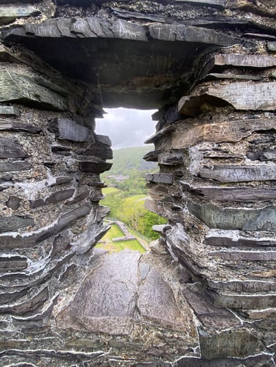 A low-angle, slightly distorted view from within a dark, rough-hewn stone structure, looking outward toward a bright landscape. The stone walls are composed of layered, textured gray and black stones, some marked with white streaks or carvings. The opening in the stone structure frames a green landscape with a patch of grass, trees, and stone features. In the background, a few buildings can be distinguished. The scene appears to be in Llanberis, United Kingdom.