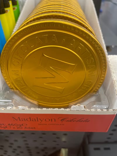 The image shows a close-up, slightly angled view of stacked gold-colored chocolate medals, likely a product packaging. The medals are circular with embossed text and a large, stylized "M" in the center. The text around the edge of the visible medals appears to read "ÇIKOLATA ARTISAN CHOCOLATE". Below the chocolate medals, a red cardboard band with white text is visible. The text on this band reads "Madalyon Çikolata" and below that, "et: 460ge", "3ge x 20 Adet", and "MEDAL CHOCOLATE". The packaging is in a white cardboard box. In the background, blurred yellow and green objects are visible, possibly other products or packaging materials. The lighting suggests an indoor environment, likely a retail setting. There is no indication of the city of Yunus Emre, Turkey within the image itself.