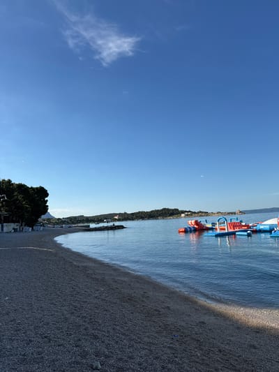 The image depicts a serene coastal scene on a sunny day, likely in Makarska, Croatia. A pebble beach curves along the left side of the frame, meeting the calm, blue sea. On the beach, several lounge chairs are visible, some with umbrellas, suggesting a resort or public area. Dense green trees line the edge of the beach, casting shadows.

In the water, a vibrant inflatable water park is set up, featuring red, blue, and orange elements, including slides and platforms. The park is positioned to the right of the image, with several people engaged in activities on or around it.

Across the water, a peninsula or island is visible, covered in lush green vegetation and dotted with buildings, including a prominent lighthouse-like structure. The sky is a clear, bright blue with a few wispy clouds, indicative of good weather. The overall impression is one of a tranquil vacation destination.