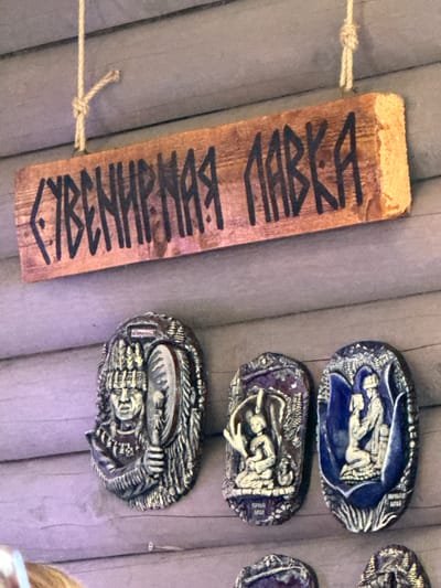 This is a medium shot, eye-level photograph taken outdoors on a wooden wall. The main subject is a wooden sign with Cyrillic lettering that reads "СУВЕНИРНАЯ ЛАВКА," meaning "Souvenir Shop." The sign is weathered and has a rough texture, with black painted lettering. It is suspended by a thin rope tied in a knot.

Below the sign, three oval-shaped decorative plaques are mounted on the wall. The leftmost plaque is a relief carving of a person wearing a headdress and holding a round object. The middle and rightmost plaques feature smaller relief carvings of figures in various poses, with intricate backgrounds. The plaque on the far right is colored blue.

The wooden wall is constructed from horizontal logs. The overall lighting suggests it is daytime, possibly overcast. The photograph appears to be taken in the vicinity of Kupchegen, Russia, as indicated by the presence of souvenir items often found in tourist locations. There are no people clearly visible in the frame.