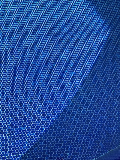The image is an abstract, top-down view of a blue tiled surface. The tiles are hexagonal and arranged in a repeating pattern. There is a diagonal gradient of light and shadow across the surface, with the upper left portion appearing brighter and the lower right portion darker. The scene is likely an outdoor tiled area, such as a pool or patio. There are no people or specific objects clearly identifiable within the frame that would indicate a precise location within Süleymanpaşa, Turkey. The focus is entirely on the texture and color of the tiled surface.