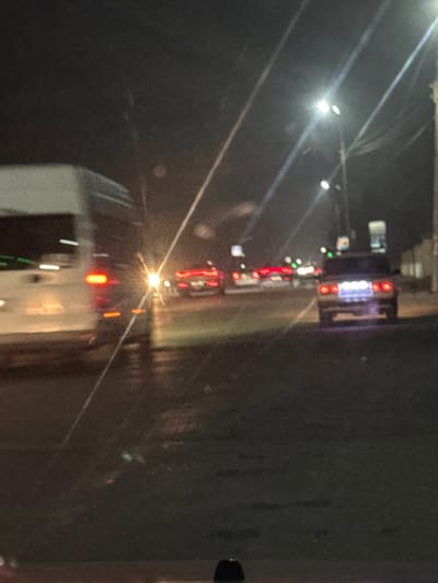 A nighttime scene shows a line of vehicles on a wet road, their headlights and taillights creating streaks of light. A large truck with illuminated signage is visible at the front of the line. The road surface reflects the lights, enhancing the visibility of lane markers. Streetlights are present, adding to the overall illumination of the area.