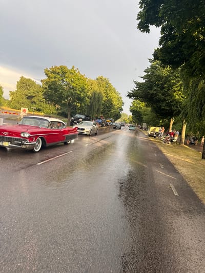 The image depicts a street scene in Falkenberg, Sweden, likely during a classic car event. A red vintage Cadillac is prominent in the foreground, driving on a wet asphalt road. Several other classic and modern cars are visible further down the road, along with a group of people gathered on the grassy verge to the right. The weather appears to be overcast, with trees lining both sides of the street. The presence of what looks like a barricade and signage suggests a designated event area. The road surface indicates recent rainfall, creating reflections from the sky and surrounding lights.