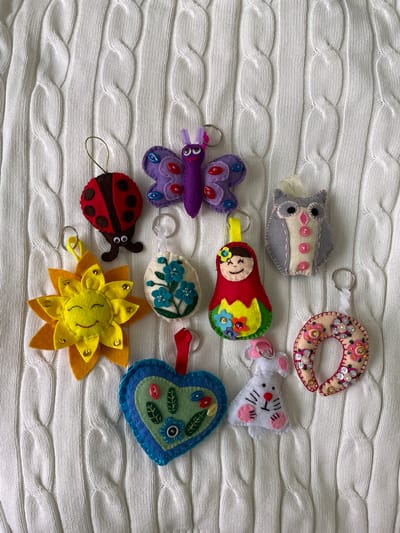 The image shows a collection of handcrafted felt keychains arranged on a white knitted fabric with a cable knit pattern. The keychains are various shapes and depict different themes, including an owl, a donut, a mouse, a butterfly, a matryoshka doll, an egg with flowers, a heart, a ladybug, and a sunflower. Each keychain has a metal keyring attached. The background is a textured white fabric, and the scene is captured in a well-lit, overhead shot. The city of Лесной Пейзаж Кп (шарапово, Russia is not depicted or implied in the visual information.