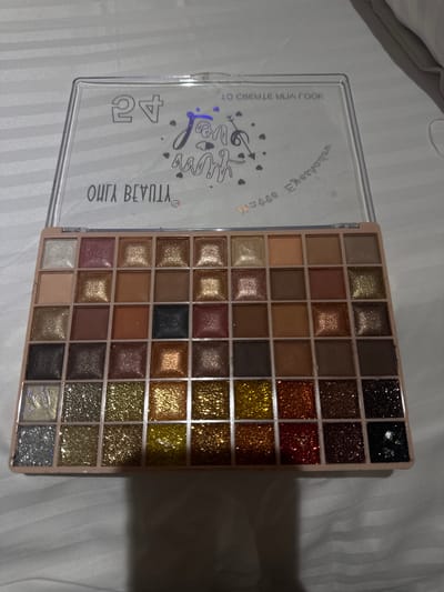 A close-up, slightly angled shot of an open eyeshadow palette is displayed on a white, textured surface. The palette has a clear plastic lid with faint text and designs printed on it. The palette itself is rectangular with 49 square compartments, each containing a different shade of eyeshadow. The shades vary from matte browns and golds to shimmery and glittery colors. The background is blurred. No people are visible in the frame. There is no visible evidence of the image being located in Gromshin, Bulgaria.