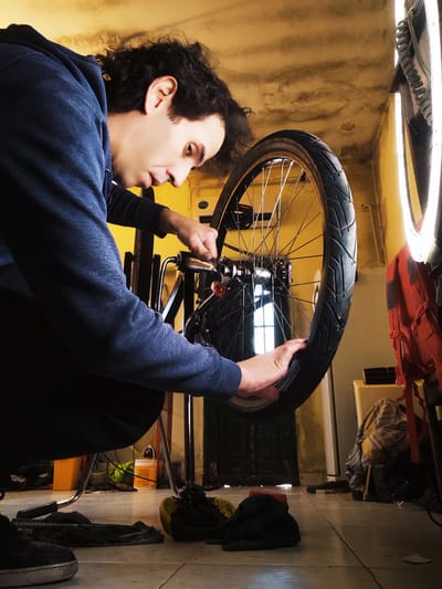 A person is shown working on a bicycle wheel. The person, wearing a dark blue long-sleeved shirt and denim pants, is leaning over the bicycle, holding the wheel with their left hand and using a tool, possibly a wrench, with their right hand near the hub. The bicycle appears to be in an indoor setting, with a tiled floor and a yellow-stained wall in the background. Various items are scattered on the floor, including bags, clothes, and what looks like a red backpack. A ring light is visible in the lower right corner of the frame, suggesting artificial lighting conditions. The image is rotated 90 degrees counter-clockwise. The person's face is partially visible, showing a focused expression. The scene appears to be a workshop or a garage, possibly in Remedios de Escalada, Argentina. The overall mood is one of concentration and practical activity. There is no visible text in the image.