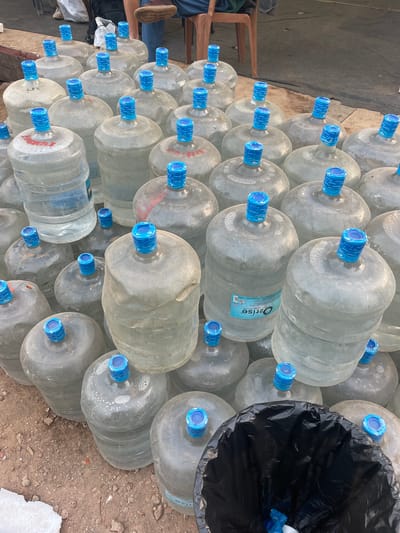 The image depicts a large stack of approximately 50 empty, transparent, reusable water bottles with blue caps. The bottles, possibly 5-gallon size, are arranged in a haphazard, multi-layered pyramid on the ground, with some bottles bearing a label reading "Ozrisse." A black trash bag is partially visible on the right side of the stack, seemingly containing some of the discarded bottle caps. The bottles are resting on an uneven, dirt or gravel-covered surface, possibly outdoors. Parts of a human being in a chair are visible in the frame. The scene is presumed to be in Mira-Bhayander, India.