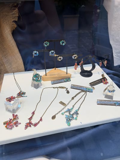 This image depicts a jewelry display case, likely from a store in Würzburg, Germany. The display is set against a dark blue fabric background, with a textured burlap material visible on the left. The case contains a variety of exquisite handcrafted jewelry pieces, including earrings, necklaces, and rings. These items are meticulously arranged on white surfaces and black stands, showcasing their intricate designs and sparkling gemstones. Notably, several pieces are branded with the name "KONPLOTT," indicating the designer or brand. The lighting in the display case appears to be focused on highlighting the jewelry, creating a polished and inviting presentation. No individuals are visible in the frame, suggesting a retail environment focused on product display.