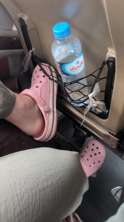 The image shows the interior of a vehicle, likely a car or bus, focusing on the legs and feet of passengers. A bottle of water with a blue cap rests in a black net pocket attached to the back of a seat. One person is wearing pink Crocs and grey pants, with their left foot visible. Another person's leg, clad in light-colored trousers and also wearing pink Crocs, is in the foreground. The upholstery of the vehicle appears to be beige. The scene suggests a journey, possibly within the city of Somsavan, Laos.