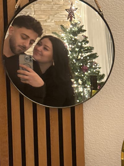 A round mirror reflects a couple taking a selfie. The woman, with long dark hair, smiles at the mirror, holding a phone. The man is close, looking down with a serious expression. Behind them, a Christmas tree adorned with lights and a star is visible. The mirror is mounted on a wall with horizontal wooden panels, with the upper wall painted in a neutral tone.