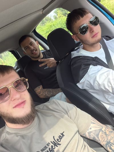 The image, rotated 90 degrees counter-clockwise, captures three young adult men inside a car during what appears to be a casual, daytime moment. Bright green foliage outside the windows suggests an outdoor setting, possibly rural or suburban. The image was captured in Vresovo, Bulgaria.

In the foreground, on the bottom right, a man with a light beard wears pink-tinted aviator sunglasses. He is dressed in a light grey/beige sweatshirt, on which the upside-down text "MADE BY" and "Wear" is visible. His right arm, featuring a detailed sleeve tattoo of playing cards, a clock, and roses, is also prominent.

To his left, towards the top of the frame, another man with short hair and aviator sunglasses is seated. He wears a white t-shirt underneath a black and white jacket. Both these men are looking towards the camera.

Further back in the car, on the middle-left, a third man with a short beard and dark hair is seen. He wears a black long-sleeved shirt with the text "ICONA CO." visible on the chest. He smiles subtly, with his tongue slightly out, and makes a peace sign with his right hand. A tattoo sleeve is also visible on his arm.

The car's interior features black seats with white stitching and a light grey roof lining. All three men appear relaxed and in good spirits.