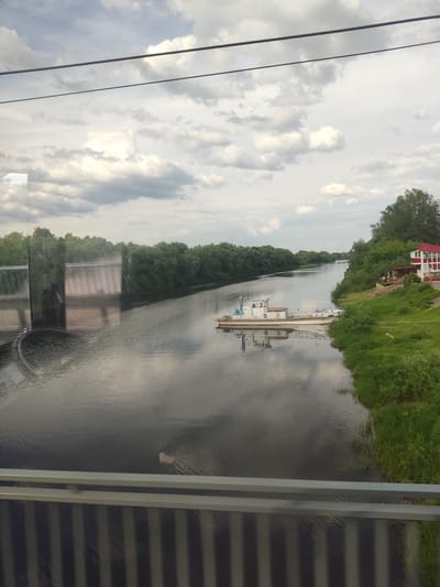 This detailed image captures a daytime riverine landscape within the city of Staraya Russa, Russia, viewed from an elevated position, likely a bridge or an observation point through a reflective glass barrier.

The scene is dominated by a dark, calm river that flows from the background towards the foreground, reflecting the overcast sky and elements of the surrounding environment. The water surface exhibits subtle ripples, causing slight distortion in the reflections.

The sky occupies the upper portion of the frame, characterized by a dense cover of predominantly grey and white cumulus clouds, indicating an overcast or partly cloudy day. Two horizontal parallel lines, possibly power lines or structural elements of a bridge, traverse the uppermost section of the sky.

The left bank of the river is lined with a dense expanse of lush green deciduous trees, extending into the distant background. In the mid-left foreground, a substantial concrete or metal structure, possibly part of an embankment or a weir, is partially visible. Reflections on the immediate foreground glass obscure some of the details of this structure, but it appears as a large, grey, vertical wall, with a dark, indistinct reflection of a person (likely the photographer) overlaid on it.

On the right bank, a white motorboat or small passenger vessel is moored parallel to the shore. The boat features a cabin structure, railings, and blue accents, with its clear reflection visible in the river's surface. The riverbank immediately behind the boat slopes upwards, covered in vibrant green grass and scattered smaller shrubs. Further up the bank, a building with a distinctive red gable roof and white walls is situated amidst the greenery. An outdoor terrace or seating area is visible adjacent to the building. A large, mature tree stands behind and to the right of this structure, with more dense green trees lining the bank into the distance.

The immediate foreground features a grey metal railing or frame, consisting of two prominent horizontal bars at the top and a series of vertical slats below, partially obscuring the lower river and bank. The reflective surface through which the photograph was taken shows various distorted reflections, including the aforementioned silhouette on the left. No people are discernible in the main landscape itself.