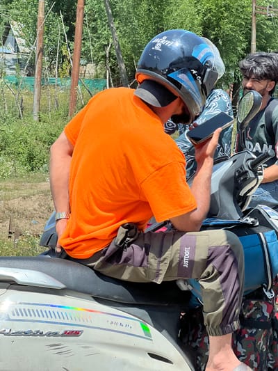 In Ujroo, India, two men are on a scooter outdoors. The man in the foreground, wearing an orange t-shirt and a helmet, is holding a smartphone. He is seated on the scooter and has a watch on his left wrist. The second man, partially visible, wears a dark t-shirt with a graphic print and is looking towards the first man. They are positioned on a scooter with a silver body. The environment is rural, with trees, vegetation, and wooden poles in the background. The lighting suggests it is daytime.