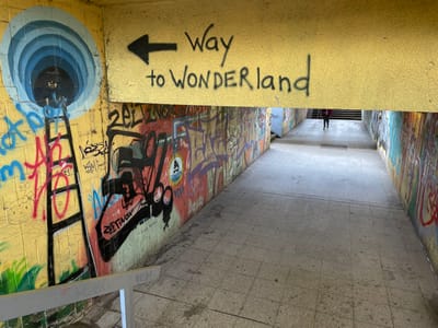 The image depicts a graffiti-covered pedestrian underpass in Oberhausen, Germany, specifically located near the Gasometer Oberhausen and OLGA Park area. The underpass walls are extensively adorned with multiple layers of colorful graffiti and tags. The prominent left wall features a yellow background with a large blue circular tunnel graphic, from which a black ladder extends downwards. Above this artwork, on the yellow overhead beam, a black arrow points left, accompanied by the spray-painted text "Way to WONDERland." The right wall is similarly covered in vibrant graffiti, showcasing various designs and lettering. The floor is constructed of light grey square tiles, showing signs of wear and dirt. In the far distance, a solitary individual, dressed in a dark jacket or top and dark trousers, walks away from the viewer towards a visible set of concrete stairs leading out of the underpass. A light grey metal railing is present in the lower left foreground, indicating a transition in elevation or a small set of steps. The underpass is illuminated by a combination of artificial lighting and ambient daylight from the exits.