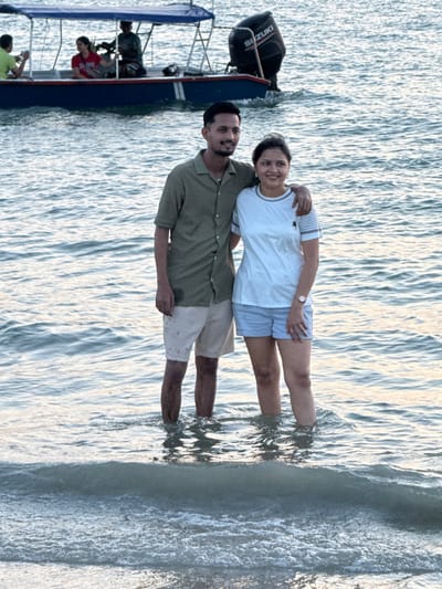 Two individuals, a male and a female, are standing in the shallow waters of Cenang Beach, Malaysia, posing for a photograph. The water reaches mid-calf level on both subjects. The male, positioned on the left, wears an olive-green short-sleeved collared shirt and light-colored shorts; his left arm is around the female's shoulders. He has short dark hair, a slight beard, and smiles towards the camera. The female, on the right, wears a white short-sleeved t-shirt with striped trim on the collar and sleeves, and light blue shorts. She has a watch on her left wrist and smiles towards the camera. In the background, a blue motorboat with a canopy and a black "SUZUKI" outboard motor is visible further out in the water. At least three other individuals are present on the boat: one in a light green shirt, one in a red shirt, and another partially obscured figure. The water is calm with gentle ripples, and the lighting is soft.
