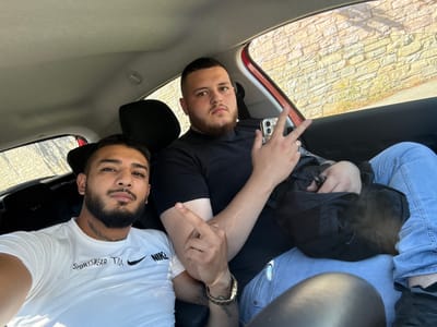 An upside-down, eye-level shot captures two men inside a car, seemingly reclining or lying down, looking towards the camera. The image is taken from inside the vehicle, with the exterior visible through the side windows.

The man on the right (left in the rotated image) wears a white t-shirt with "NIKE" and "SPORTWEAR TM" text, light blue jeans, and a gold wristwatch. He has a beard, mustache, and visible tattoos on his right arm and neck (a laurel wreath design). His right hand is raised, making an upward-pointing middle finger gesture. His expression is neutral.

The man on the left (right in the rotated image) wears a black t-shirt and light blue jeans. He also has a beard and mustache. His left hand is raised, holding a smartphone, and his fingers are positioned in a "peace" or "V" sign. His expression is also neutral.

Behind the men, parts of the car interior, including headrests and the ceiling, are visible. Through the car window, a light-colored stone or brick wall, topped with some green vegetation, is seen, indicating an outdoor setting. Bright sunlight illuminates the wall, suggesting it is daytime. A black jacket or bag is partially visible near the man in the black t-shirt.

The location context for this media file is Cocklake, United Kingdom.