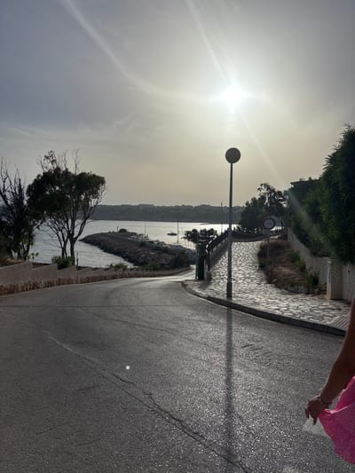A view of a coastal road and walkway in Urbanización Cabo Roig, Spain, on a sunny day. The sun is bright, positioned high in the hazy sky, casting long shadows. In the foreground, a dark asphalt road curves to the left, with faint cracks visible. A person, partially visible on the right edge of the frame, wears a pink dress with white polka dots and bracelets on their wrist. A pathway made of flagstones runs alongside the road, leading towards a harbor. A lamppost stands on the paved walkway. Beyond the walkway, a rocky breakwater extends into the sea, where several sailboats and a larger boat are moored. Trees with sparse foliage line the left side of the road and the edge of the water. In the distance, buildings are visible along the coastline. A sign with a bicycle symbol and "EXCEPTO POLICIA" (Except Police) is posted on the right side of the paved walkway.