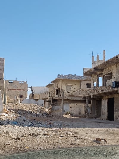 This image portrays a severely damaged urban streetscape within Al-Qadam Municipality, Syria, under a clear blue sky. The scene is characterized by multiple multi-story buildings, all exhibiting extensive structural degradation consistent with sustained conflict or demolition. Building facades are heavily pockmarked with numerous circular impact holes, suggesting gunfire or shrapnel damage. Windows and doorways are predominantly empty apertures, with some revealing exposed rebar and interior debris. Portions of concrete roofs and floors are either missing or collapsed. The ground surface is unpaved, consisting of a dense accumulation of light-colored rubble, including shattered concrete, broken bricks, and dust, forming substantial piles in the foreground and mid-ground. No individuals, vehicles, or active elements are discernible within the frame, indicating a deserted and dilapidated environment.