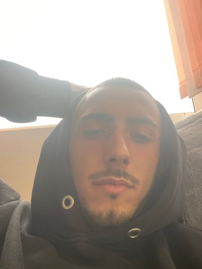 The image presents a close-up, frontal, selfie-style photograph of a young adult male, taken indoors in a location stated to be Salmanovo, Bulgaria. The subject has short, dark hair, a mustache, and a short beard. He is wearing a dark, possibly black, hooded sweatshirt; two metallic eyelets are visible on the lower front edge of the hood. A light-skinned hand, identifiable as belonging to a second individual, is visible in the upper left quadrant, resting on the back of the primary subject's head. The background is heavily overexposed due to intense light, consistent with a bright window. Partially visible on the upper right are vertical blinds featuring alternating light orange/brown and cream stripes. A textured, light-grey fabric object, consistent with a blanket or pillow, is discernible behind the subject's right shoulder (viewer's left).