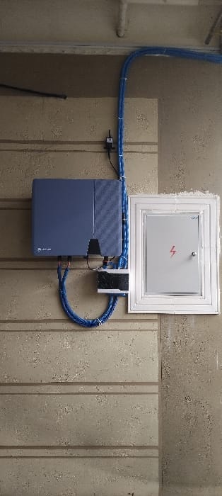 A blue battery unit is mounted on a wall with a metal-framed access panel above. The panel features a red lightning bolt symbol. Blue cables connect the battery to the panel and a black device to the right. The wall is textured with alternating light and dark sections, with a lighter section behind the battery and panel. A pipe and cables are visible on the left side of the wall.