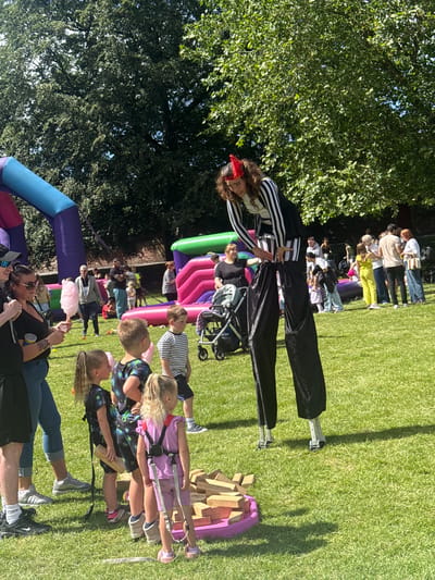 The image depicts a sunny outdoor event, likely a fair or festival, taking place on a grassy lawn at the Pinsley Green Leisure Centre in Leominster, United Kingdom. The central focus is a performer on stilts, dressed in a black and white striped top and baggy black pants, wearing a red headpiece. They are interacting with a group of children, one of whom is holding a pink cotton candy. In the background, several inflatable structures are visible, including a large purple and blue bouncy castle on the left and a pink and green one on the right. Various people, including adults and children, are scattered across the lawn, some watching the stilt walker, others conversing or attending to children. A few children are gathered around a low platform with large wooden blocks, suggesting a game or activity. The environment is characterized by lush green grass and mature trees providing shade. The overall scene conveys a sense of lively community engagement and family-friendly entertainment.