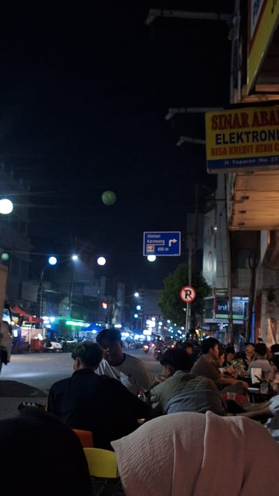 The image shows a street scene at night in Karawang, Indonesia. A sign indicates that the Karawang train station is 450 meters away. Another sign advertises "Sinar Abadi Electronics" at Jl. Tuparev No. 277. People are seated, possibly at a restaurant, and several motorcycles are visible in the background. The illumination comes from streetlights and the lights of various businesses. A no-right-turn sign is also present.