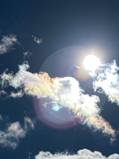The image depicts a deep blue sky with scattered clouds. The sun is visible, appearing as a bright white orb, creating a lens flare and halo effect. Some of the clouds exhibit iridescent colors, suggesting they might be nacreous clouds or showing diffraction effects. The scene is devoid of any man-made structures or people, making it impossible to determine a specific location within the city of Gela, Bulgaria, or any other city. The focus is solely on the atmospheric phenomena.