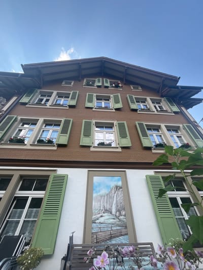 The image displays the facade of a multi-story building located in Lauterbrunnen, Switzerland, captured from a low-angle perspective against a blue sky with scattered white clouds.
The building's upper three levels are characterized by a brown, scaled, shingled exterior and a prominent, dark-eaved gabled roof. The two main upper floors each contain three rectangular windows with white frames and open, light-green wooden shutters. Most of these windows feature window boxes with red flowers. The uppermost, attic level presents two smaller windows, one of which also displays red flowers in a window box.
The ground floor of the building is distinguished by a white plastered facade. It features two large, rectangular windows with white frames. The left ground-floor window has one light-green shutter open and another partially closed. Between these two windows, a vertical, framed mural is painted directly onto the white wall. The mural depicts a scenic landscape with a multi-tiered waterfall cascading down a grey cliff face, a small, white-roofed structure at its base, and a wooden arch bridge spanning a river in the foreground of the painting.
In the immediate foreground, to the left, a portion of a dark-colored outdoor chair and small white potted flowers are visible. Towards the bottom center, a cluster of light purple and pink flowers with yellow centers is present, in front of a partially visible wooden slatted bench. On the right side of the foreground, large green leaves from a plant extend into the frame, partially obscuring the right ground-floor window. No individuals are depicted in the scene.