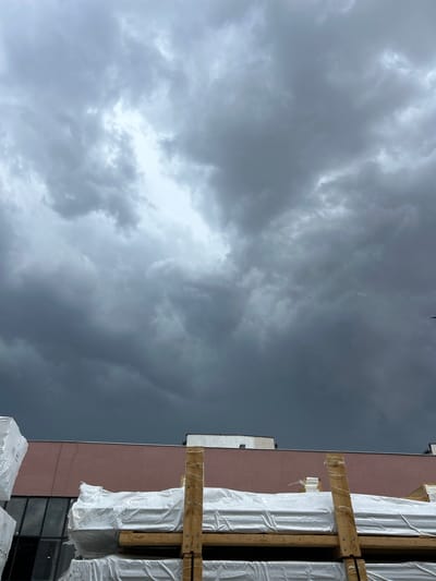 The image shows a view of a building with a pink facade, large windows, and a white roofline. In front of the building, there are stacks of white-wrapped materials on wooden pallets. The sky is overcast with dark, heavy clouds. The scene appears to be an outdoor storage area or loading dock. There is no indication of the city of Struma, Bulgaria, in the image.