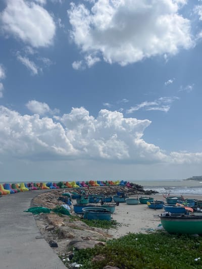 The image depicts a coastal scene in Phú Thủy, Vietnam, on a bright, partly cloudy day. A concrete pathway lines the left side of the frame, leading towards a colorful barrier of tetrapods (concrete breakwater blocks) in shades of blue, green, yellow, and orange. Beyond the tetrapods, a sandy beach is visible, cluttered with numerous blue and green basket boats, some appearing empty and others with fishing gear. The sea, with moderate waves, stretches towards the horizon, where a town or resort is faintly visible on a distant headland. The foreground shows scattered rocks and low-lying green vegetation.