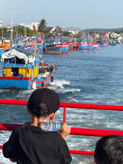 A medium shot captures a young boy wearing a black t-shirt and a black baseball cap, standing on a boat and looking out at a fleet of brightly colored fishing boats docked in the water. He holds a clear plastic water bottle in his right hand, resting it on the red railing in front of him. The fishing boats are predominantly painted in blue and red, with yellow accents and white canopies. Some of the boats have red flags flying. In the background, the shoreline is visible with buildings and palm trees, suggesting a coastal or harbor area. The water is blue and choppy, indicating movement. The lighting suggests it is daytime, with the sun casting bright reflections on the water. The location appears to be An Hai, Vietnam, given the style of the boats and the surrounding architecture.