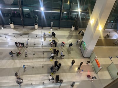 This is a high-angle, overhead photograph of an indoor airport terminal in Pajang, Indonesia, at night. The terminal features a polished, light-colored tiled floor with a grid pattern of darker lines. The ceiling is high and largely obscured by lighting fixtures and structural elements, with large windows lining the upper portions of the walls, reflecting interior lights and the dark exterior.
Several rows of stanchions with black ropes guide queues for what appear to be check-in counters or security checkpoints. People are seen scattered throughout the terminal, many with luggage. The individuals are of various ages and ethnicities, dressed in casual and professional attire.
Specific actions include:
- A group of people, some with luggage, standing in a queue at a counter labeled "1".
- A person in uniform appears to be attending to individuals at the counter.
- Another queue, labeled "3", also shows people with luggage interacting with staff.
- A person in a blue shirt is pushing a cart with luggage past a queue.
- A group of people are walking with suitcases, some are sitting on benches, and others are engaged in conversation.
- At a counter labeled "5", people are checking in or going through security.
- Further down, a "RED CHANNEL" sign with an arrow is visible, indicating a specific pathway or service.
- Some individuals are seated, possibly waiting for flights.
- Two large, cylindrical planters with dark green artificial plants are positioned near the large windows.
- A tall, angular, white pillar or structural element is prominent in the mid-right of the frame.
- Various pieces of luggage, including suitcases of different sizes and colors, are present, being pulled or carried by individuals.
- The lighting in the terminal is a mix of bright overhead lights and softer, warmer lights emanating from fixtures on the pillar.
- Reflections are visible on the floor and windows, adding to the sense of depth and space.
- Some people are wearing masks, suggesting ongoing public health protocols.
- The overall atmosphere appears busy but orderly.
