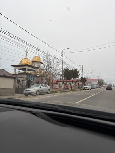 The image depicts a street scene in 2 Mai, Romania, captured from the interior of a vehicle, showing a portion of the dashboard and windshield wipers in the immediate foreground. On the left, a prominent Orthodox church structure is visible, featuring two golden, onion-shaped domes, each topped with an Orthodox cross. The church's lower exterior walls display colorful religious murals. A metal fence separates the church property from a sidewalk where a silver four-door hatchback is parked. Numerous overhead power lines and utility poles with streetlights traverse the scene. To the right, a two-lane asphalt road extends into the distance, marked with a solid white line. Several vehicles are parked along the right side of the street, including a white SUV, and additional vehicles are visible traveling away from the camera in the far distance. Various residential or commercial buildings with pitched roofs line both sides of the street. The sky is uniformly overcast and light gray. No individuals are discernible.